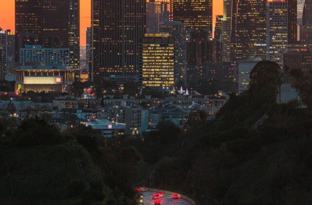 Traffic moving along a Los Angeles freeway at dusk with the city skyline behind it